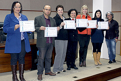 Imagem em plano geral mostra seis servidores exibindo seus certificados de conclusão do curso de Libras. Ao lado dos alunos, a professora que ministrou o curso. Todos estão olhando para a câmera, de pé, na parte da frente do auditório da Escola Judicial.