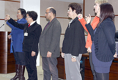 Imagem em plano geral mostra seis alunos que concluíram o curso de Libras, de pé, na parte da frente do auditório, dirigindo-se à plateia por meio de sinais.