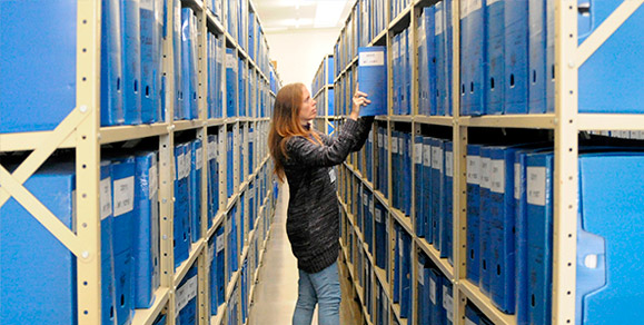 Servidora, posicionada em longo corredor com estantes de aço em ambos os lados, manuseia caixa de processos arquivados. As prateleiras das estantes estão completamente ocupadas com caixas de processos.