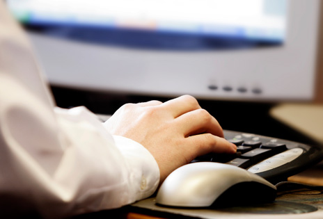 imagem fechada na mão de um trabalhador sobre um teclado, ao lado de um mouse e diante da tela do computador que aparece parcialmente ao fundo desfocada