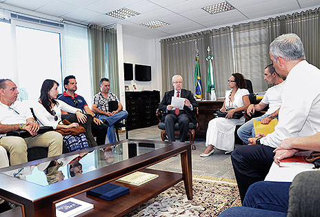 Imagem em plano aberto mostra o desembargador Altino Pedrozo dos Santos reunido em seu gabinete com oficiais de Justiça do TRT-PR e da Justiça Federal. Todos estão sentados em poltronas e cadeiras, ao redor de uma mesa de centro. O presidente do TRT-PR aparece de frente para a câmera, ao fundo, e os servidores encontram-se posicionados dos dois lados do magistrado, dispostos em semicírculo. A mesa de centro e alguns oficiais de Justiça aparecem em primeiro plano