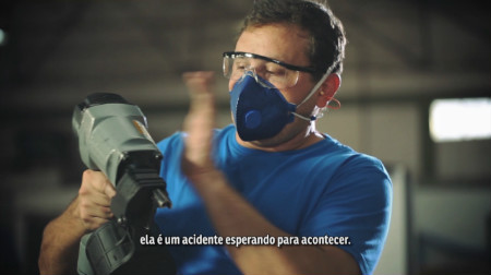 Imagem em plano médio mostra operário manuseando uma pistola de pregos. O empregado está usando óculos de proteção e máscara sobre o nariz e a boca, e está fazendo um movimento como se estivesse para bater com uma das mãos na parte lateral do equipamento. Uma legenda na imagem diz: ela é um acidente esperando para acontecer.