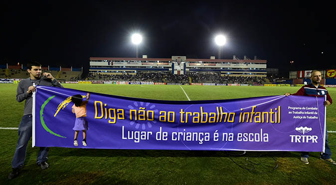 Foto em plano geral mostra faixa da campanha contra o trabalho infantil sendo exibida durante o jogo entre Paraná Clube e Luverdense. Em primeiro plano, duas pessoas seguram a faixa com os dizeres Diga não ao trabalho infantil. Lugar de criança é na escola. Ao fundo é possivel ver, parcialmente, a arquibancada do estádio Durival Britto da Silva, ocupada por torcedores.