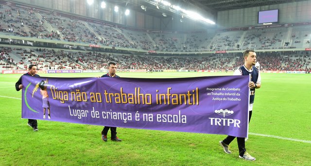 Foto em plano geral mostra faixa da campanha contra o trabalho infantil sendo exibida durante o jogo entre Atlético Paranaense e Vasco da Gama. Em primeiro plano, três pessoas conduzem a faixa com os dizeres Diga não ao trabalho infantil. Lugar de Criança é na escola. Ao fundo é possível ver parte da arquibancada do estádio Arena da Baixada, ocupada por torcedores, além de um telão