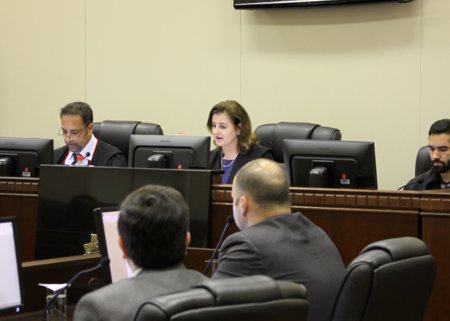 Imagem mostra parte do plenário em que foi realizada a audiência de dissídio coletivo envolvendo médicos e empresas de saúde do Paraná. Ao fundo é possível ver a desembargadora Marlene T. Fuverki Suguimatsu conduzindo a sessão. Ao seu lado estão o representante do Ministério Público do Trabalho e um assistente da magistrada. Em primeiro plano, de costas, estão dois dos representantes das empresas de saúde que estiveram presente à audiência.