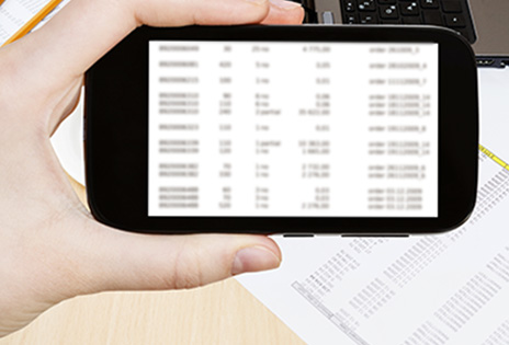 Imagem em plano fechado mostra m&atilde;o segurando telefone celular capturando imagem de documentos que aparecem sobre uma mesa, em plano mais afastado.