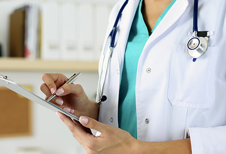 A imagem, em primeiro plano, mostra uma médica com uma prancheta à mão, escrevendo. Ainda é possível ver, no pescoço da médica, um estetoscópio.