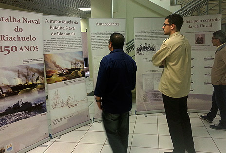 A foto, em plano geral, mostra três pessoas observando os banners da exposição.