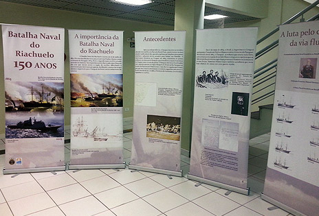 A foto, em plano geral, mostra cinco banners expostos contando a história da Batalha do Riachuelo.