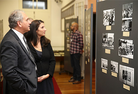 A foto, tirada em plano médio, mostra duas pessoas observando um mural com fotos de momentos históricos para os bancários. O mural está no canto direito da imagem.