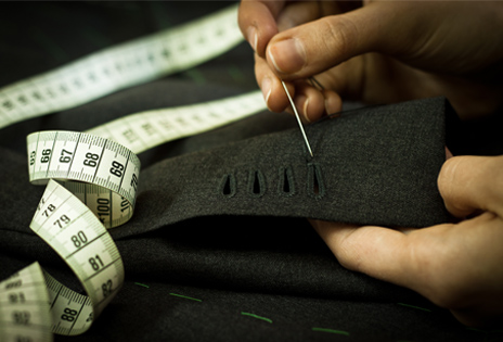 Imagem em plano fechado mostra detalhe de mãos de profissional de costura usando uma agulha na produção de peça de roupa. Sobre o tecido é possível ver parcialmente uma fita métrica típica das que se usam em oficinas de costura e alfaiatarias.